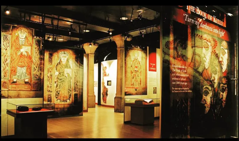 Exhibit of The Book of Kells in a museum, showcasing illuminated manuscript pages with intricate Celtic designs and religious figures. The display features warm lighting, stone pillars, and an informational panel titled 'Turning darkness into light.