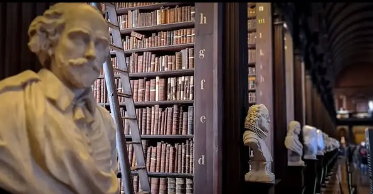 The Long Room of Trinity College Library in Dublin, featuring towering wooden bookshelves filled with ancient books, a rolling ladder, and a row of marble busts of historical figures lining the hall.